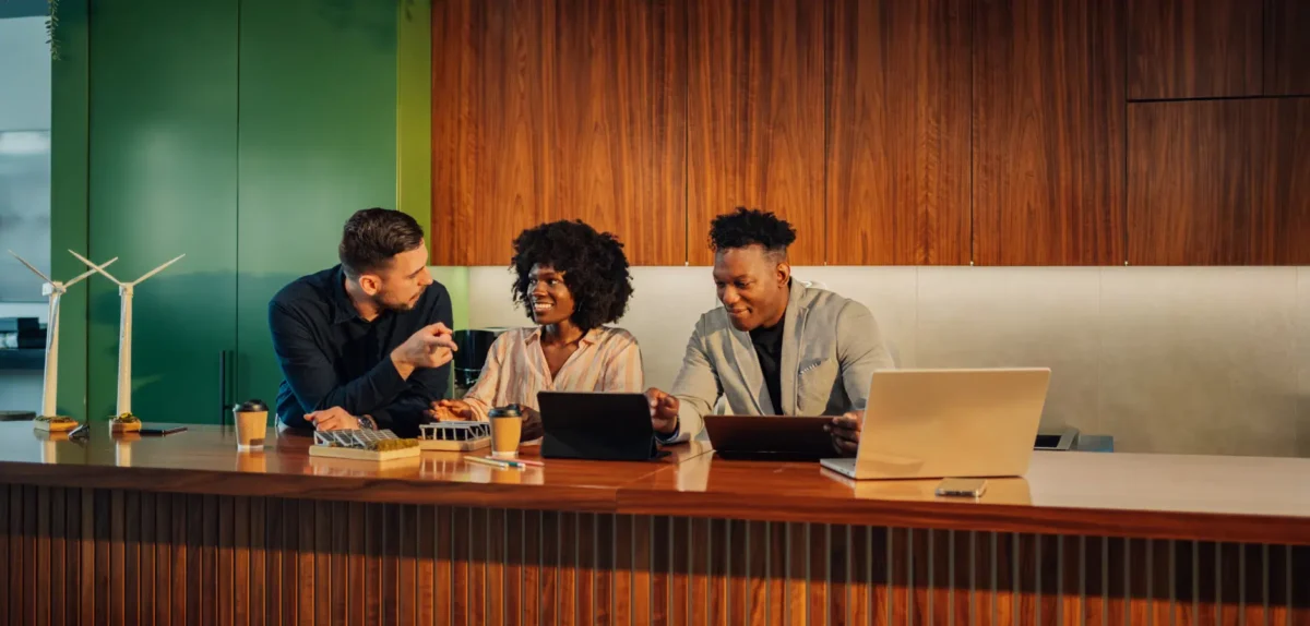 Three individuals seated at a desk, each using a laptop, engaged in collaborative work or discussion.