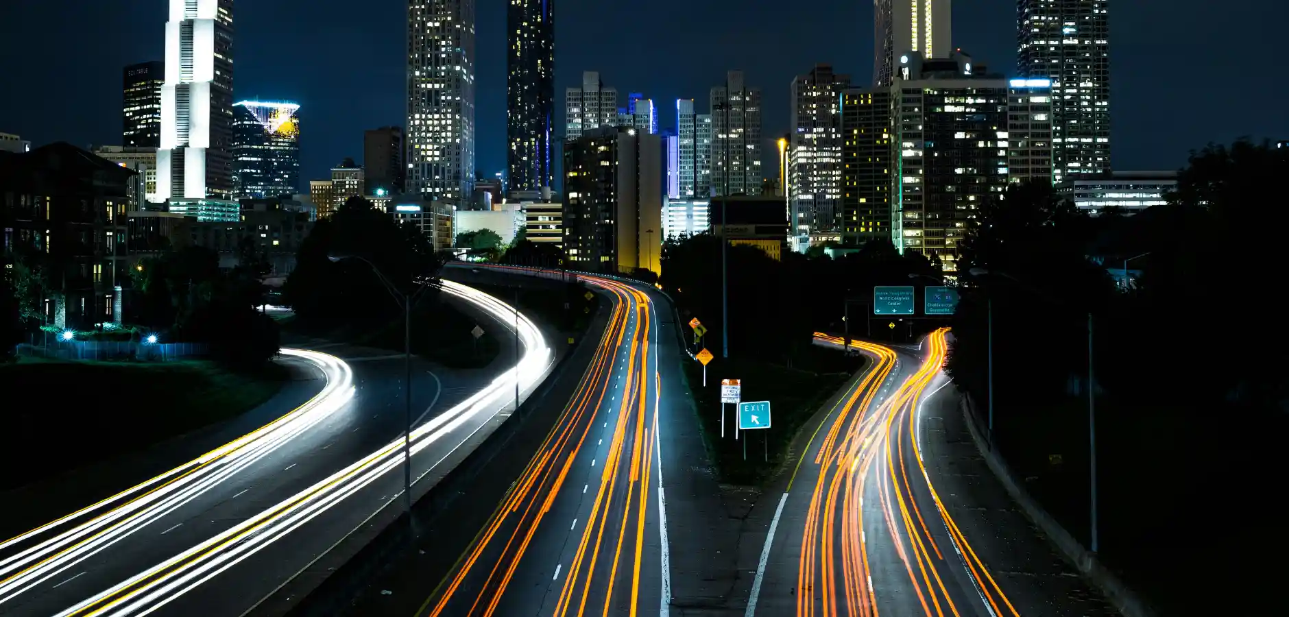 A city skyline illuminated at night, featuring a highway with streaks of light from moving cars.