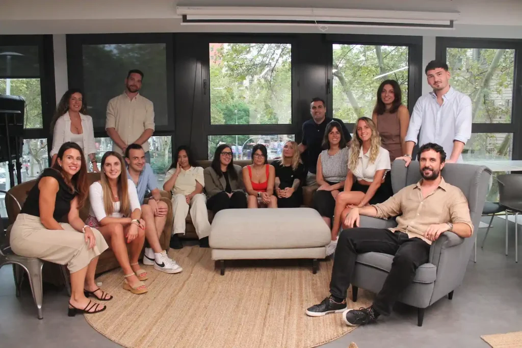 A diverse group of people gather in a modern room, seated and standing around a coffee table, with large windows providing a view outside.