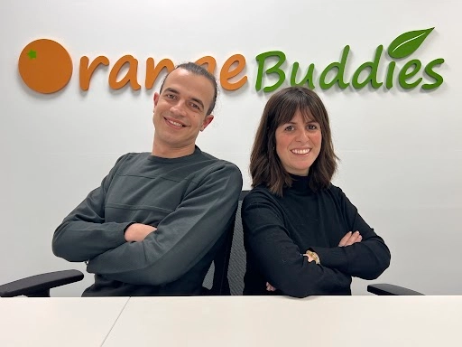 Two individuals sit back-to-back with arms crossed, in front of a wall featuring the "orangeBuddies" logo in vibrant colors.