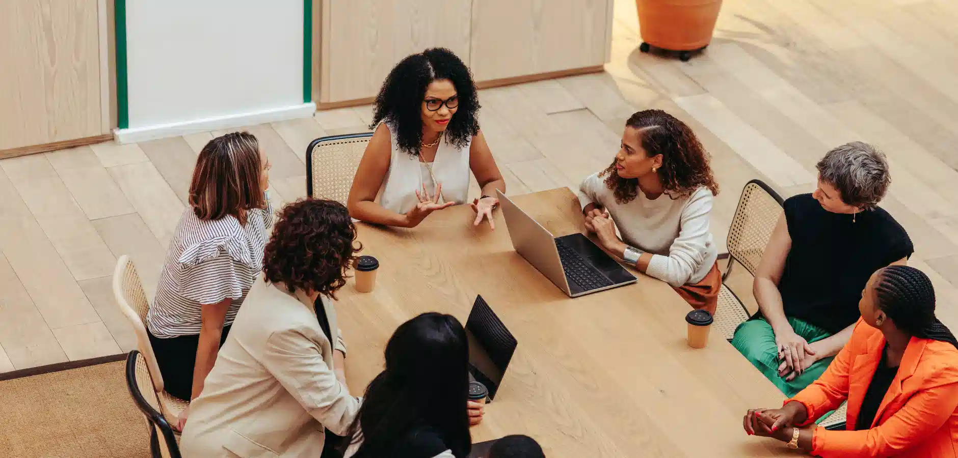 A group of diverse women engage in discussion around a wooden conference table, with laptops and coffee cups present.