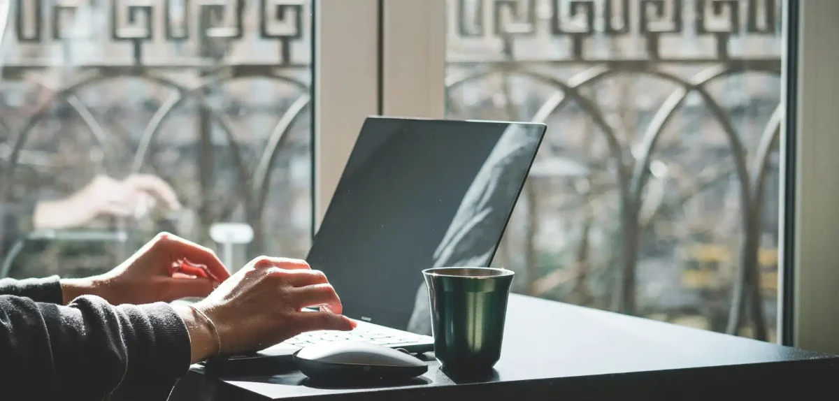 A person's hands typing on a laptop beside a green cup, viewed through a sunlit window with a decorative railing in the background.