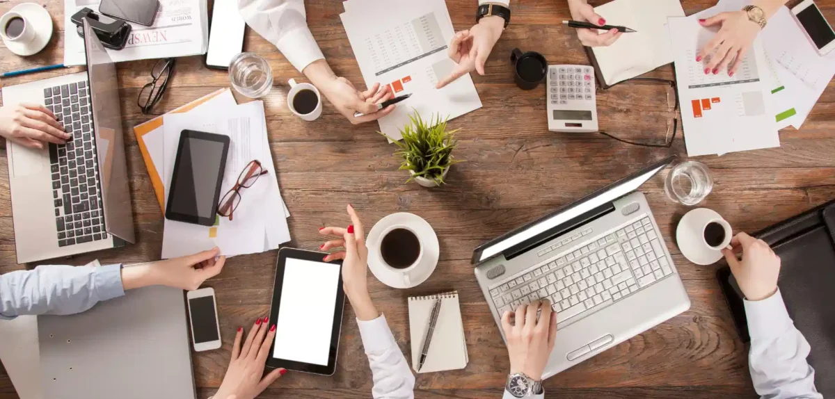 Overhead view of a busy workspace with hands holding devices, coffee, documents, and a laptop on a wooden table.