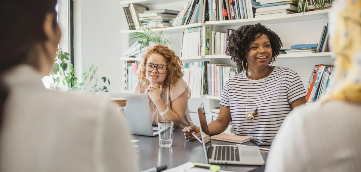 A diverse group of women engaged in a meeting at a table, with laptops and notebooks, surrounded by books and plants.