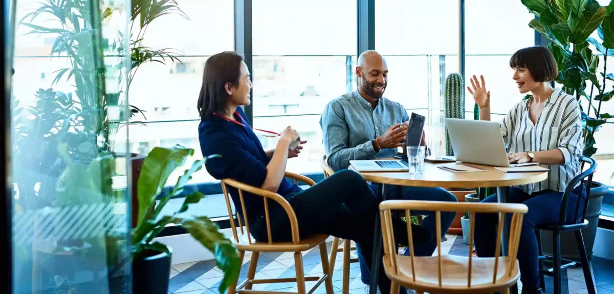 A diverse group of professionals collaborate at a sunlit table surrounded by plants, using a laptop and engaging in discussion.