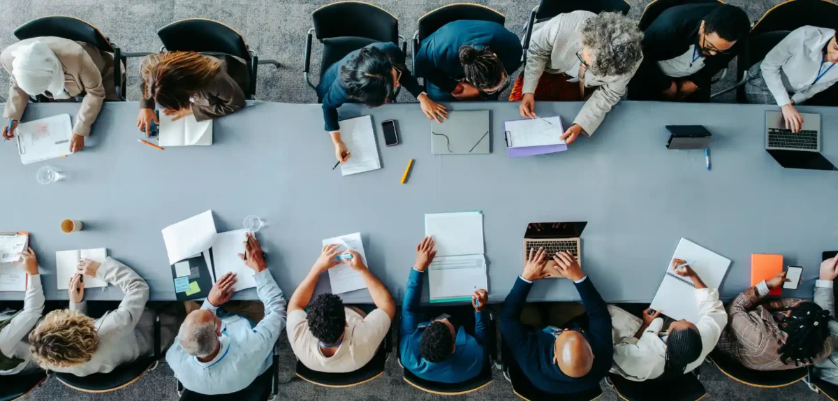 Aerial view of a diverse group of people in a meeting, seated around a lengthy table with laptops, notebooks, and stationery visible.