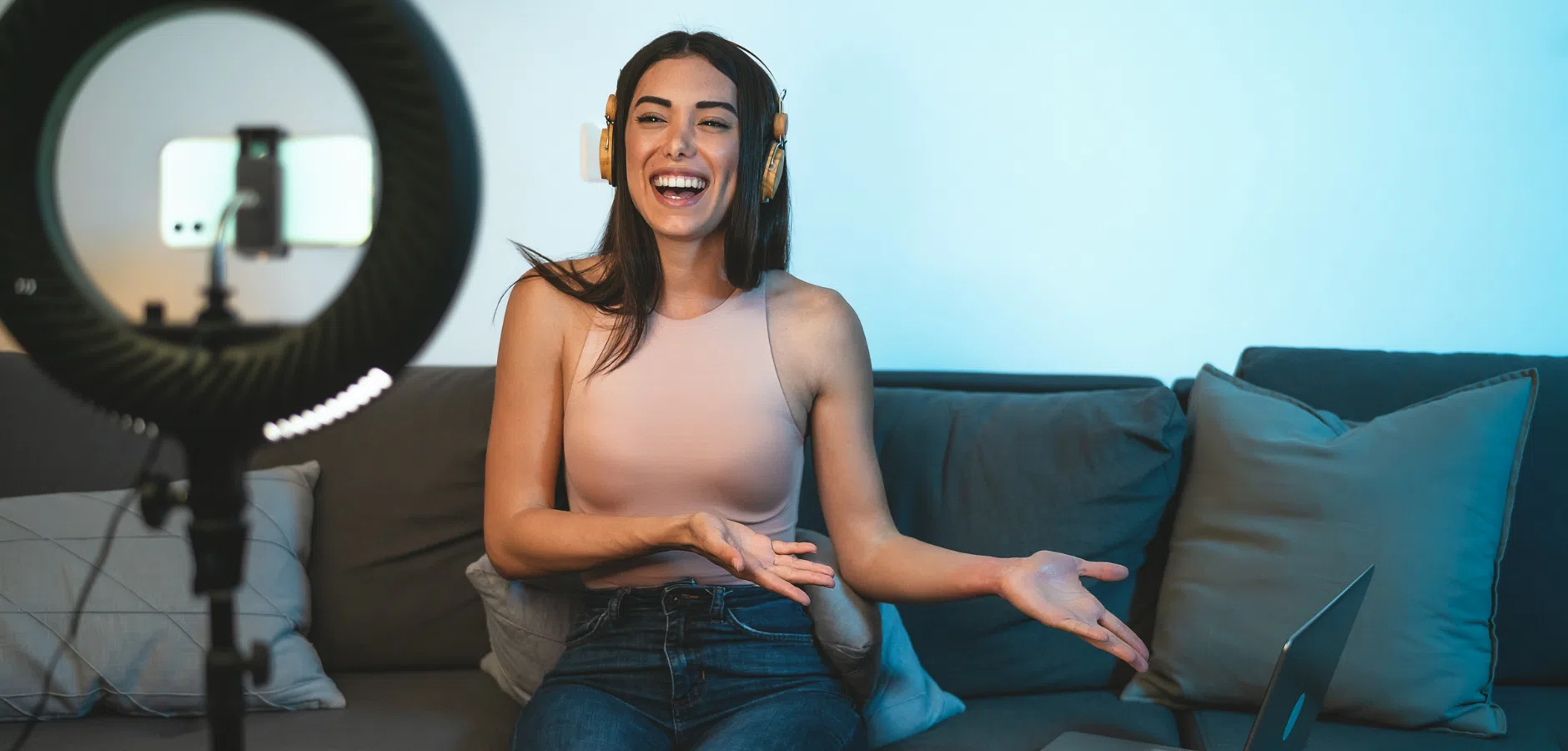 A person wearing headphones sits on a couch, gesturing while speaking into a smartphone mounted on a ring light. Laptops are nearby.