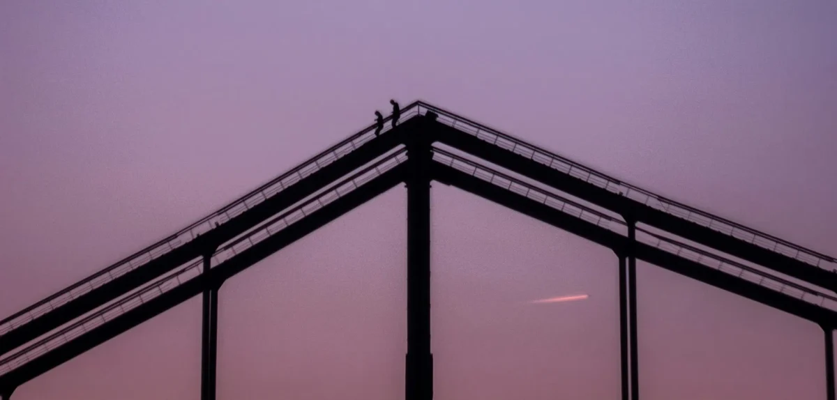 Silhouetted figures stand atop an industrial structure against a pastel purple sky at dusk.