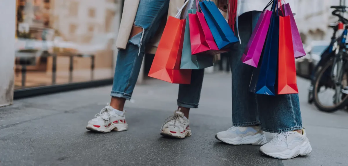 Two people stand on a city sidewalk, holding colorful shopping bags while wearing sporty shoes and casual denim outfits.