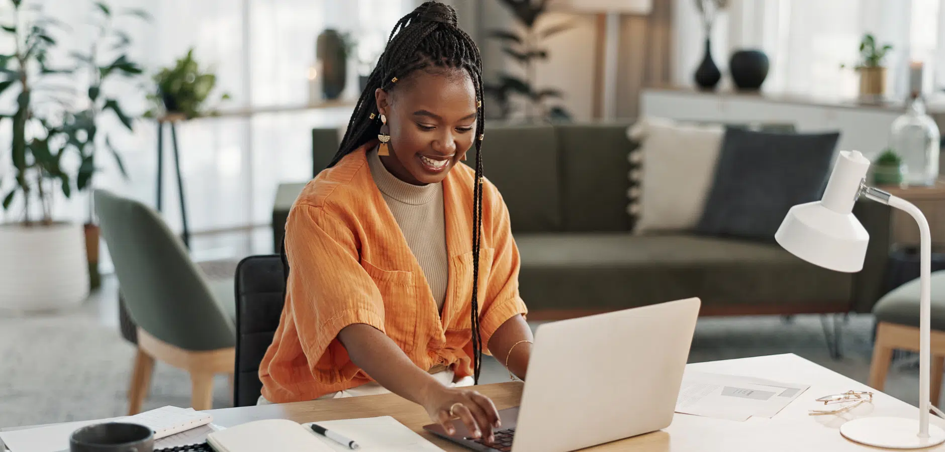 A person in an orange shirt works on a laptop at a stylish desk, surrounded by indoor plants and modern furnishings.