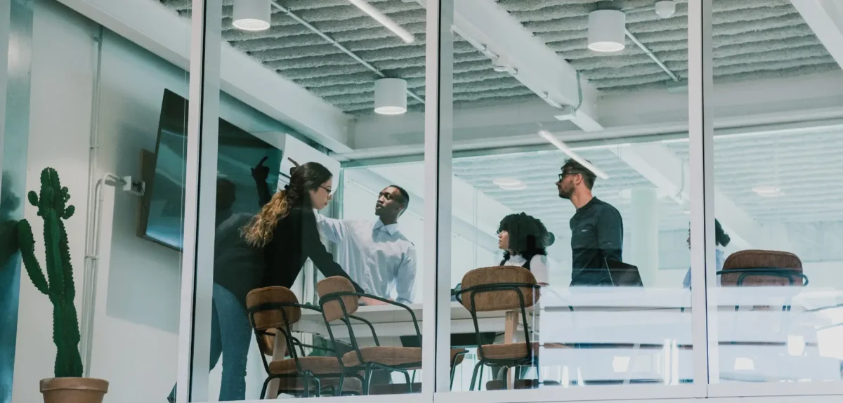 A group of professionals engages in a discussion around a conference table in a modern glass-walled meeting room. A cactus decorates the space.