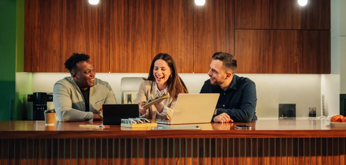 Three professionals engaged in a discussion at a modern wooden counter, with laptops and a cup of coffee in a stylish kitchen setting.