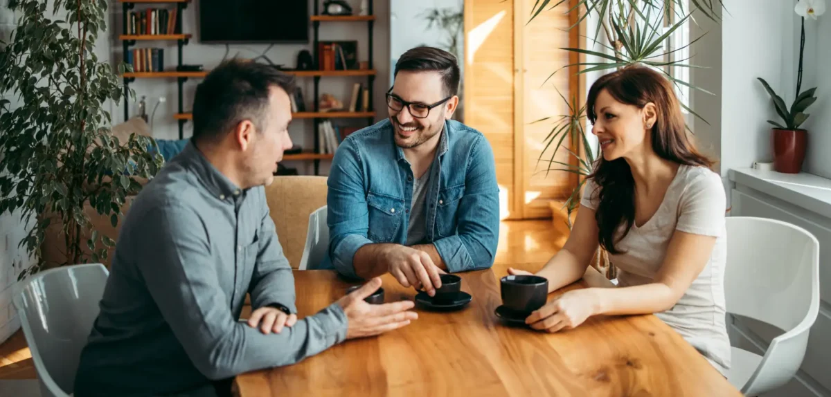 Three people are engaged in conversation around a wooden table, each holding a coffee cup in a modern, well-lit room.