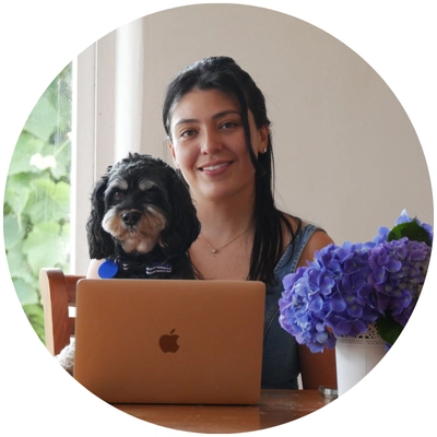 A person sits at a wooden table with a laptop, accompanied by a small dog and a vase of blue hydrangeas.