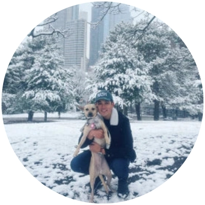 A person in a winter setting holds a tan dog, surrounded by snow-covered trees and a city skyline in the background.