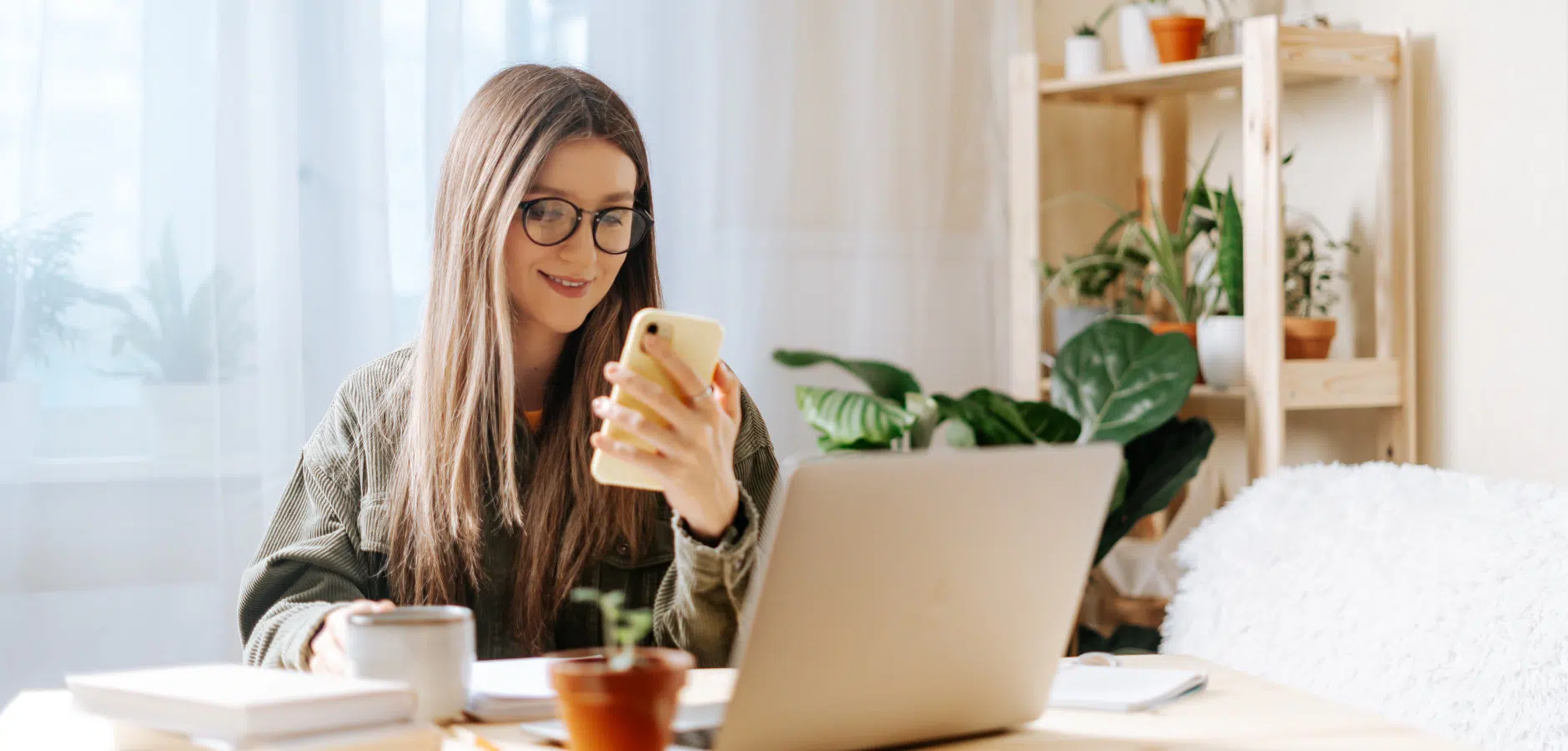 A woman sits at a desk with a laptop, holding a phone, coffee cup, and surrounded by potted plants in a bright, cozy space.