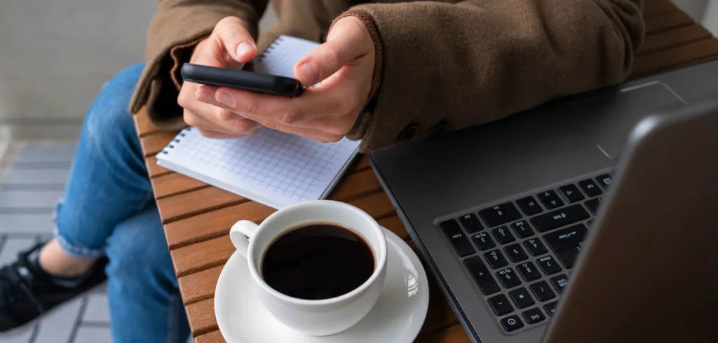 A person in a brown coat sits at a table with a laptop, coffee, and notepad, holding a smartphone and looking at the screen.
