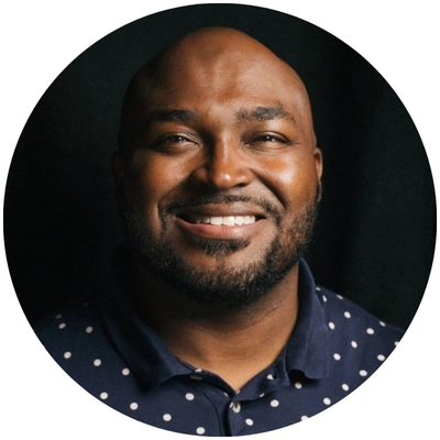 A close-up portrait of a man wearing a navy polka dot shirt, set against a dark background, with a circular frame.