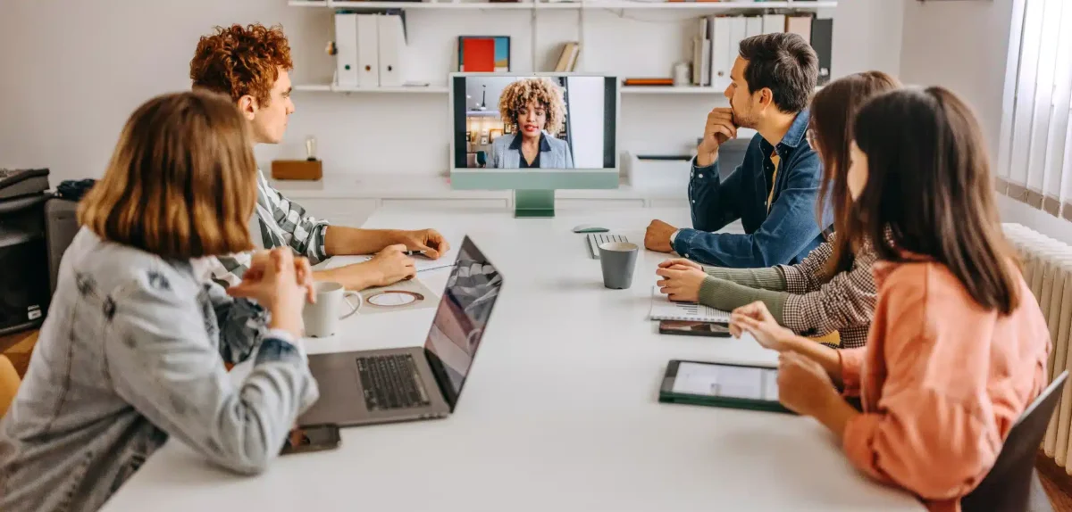 A group of six people engaged in a video call discussion in a bright, modern meeting room, with laptops and tablets on the table.