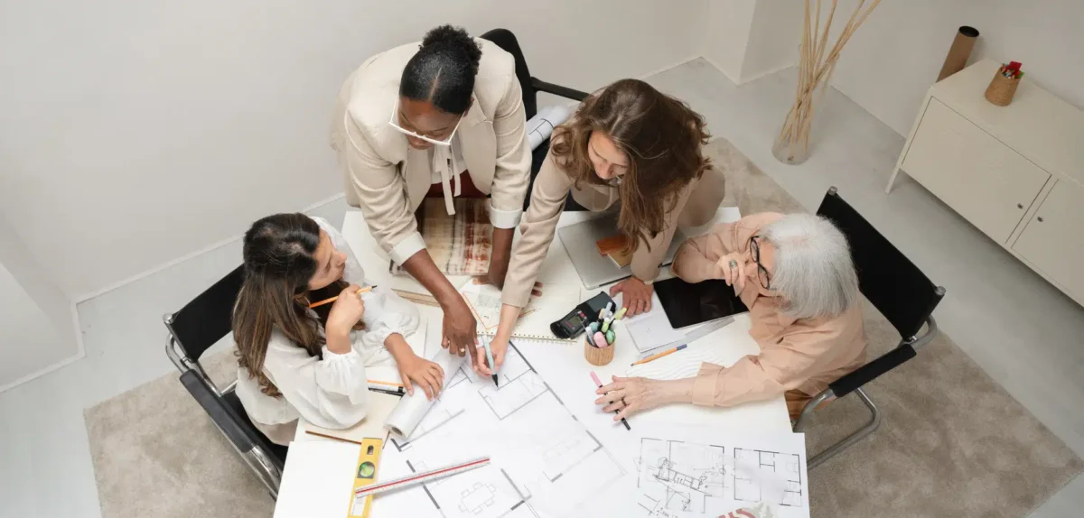 A group of diverse individuals collaborating on architectural plans at a table, surrounded by tools and notes in a bright, modern space.