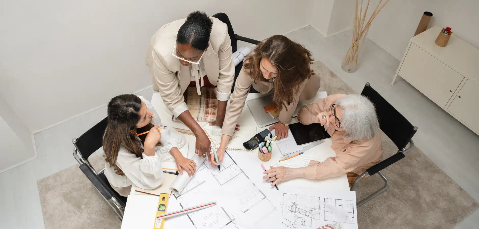 A group of diverse individuals collaborating on architectural plans at a table, surrounded by tools and notes in a bright, modern space.