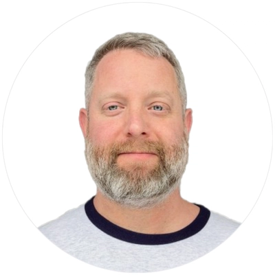 A close-up portrait of a middle-aged man with a gray beard, wearing a light gray shirt with a navy blue collar, against a white background.
