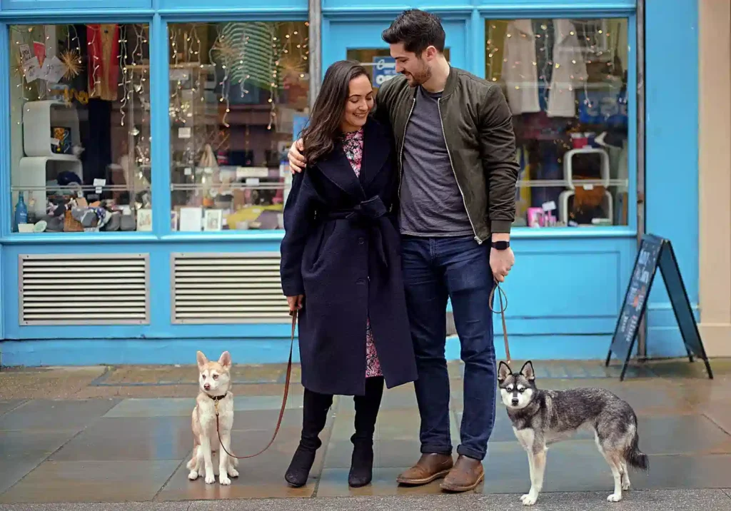 A woman in a dark coat and a man in a green jacket stand together, each holding a dog on leashes, in front of a colorful shop.