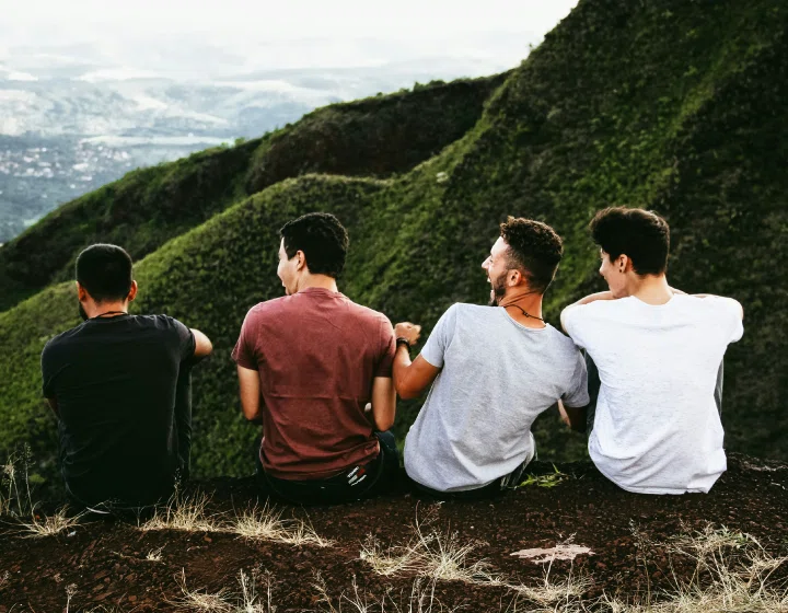 Four friends sit atop a green hillside, gazing at the distant landscape under a cloudy sky, enjoying a moment of connection and nature.