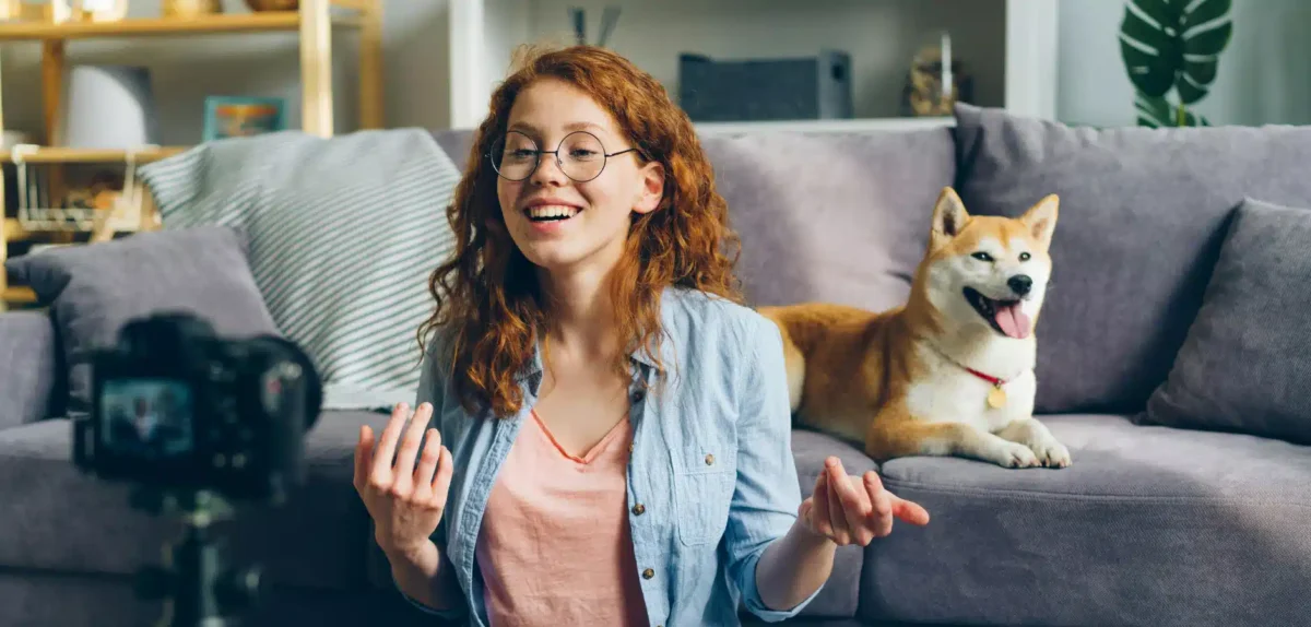 A woman sits on a couch with a dog beside her, holding a camera and smiling at the viewer.