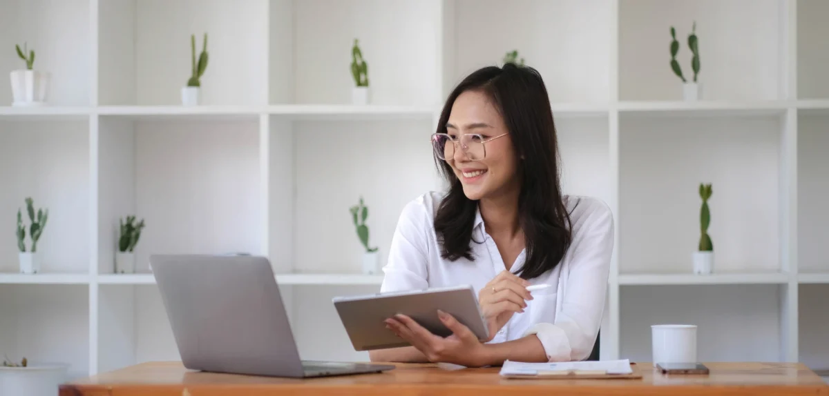 Asian woman focused on a tablet computer while sitting at a desk, surrounded by office supplies.