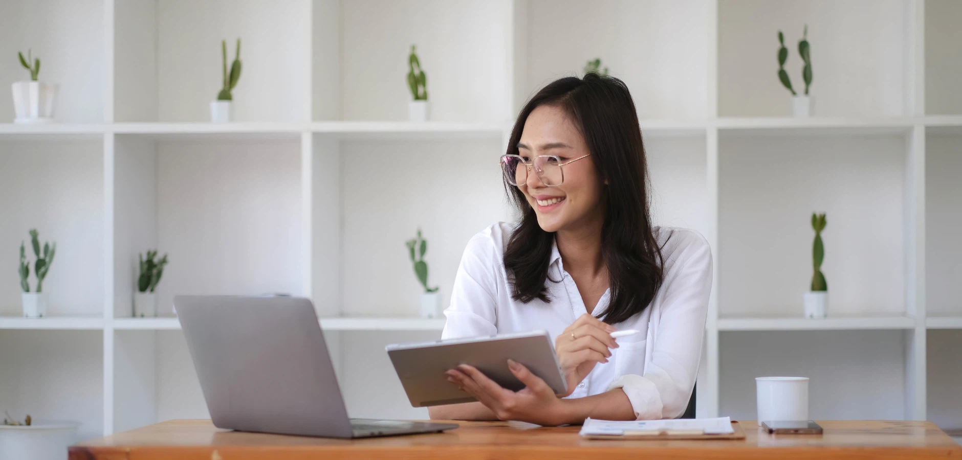 Asian woman focused on a tablet computer while sitting at a desk, surrounded by office supplies.