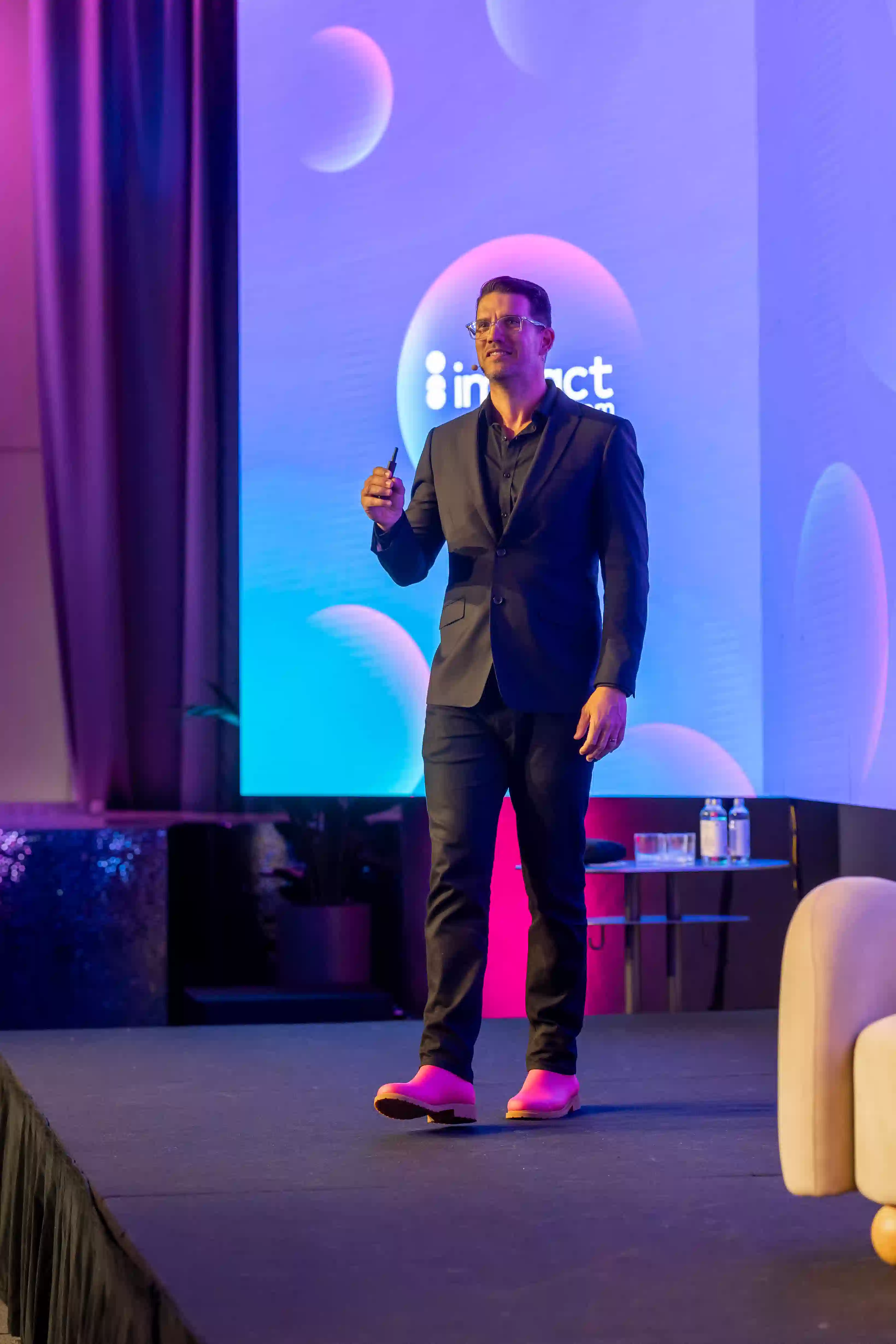 A speaker in a stylish black suit and pink shoes stands on stage, holding a pen, with a colorful backdrop featuring the word "impact."