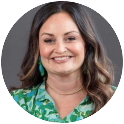 A woman with wavy, shoulder-length hair wears a vibrant green patterned blouse and a simple gold necklace, posed against a neutral background.