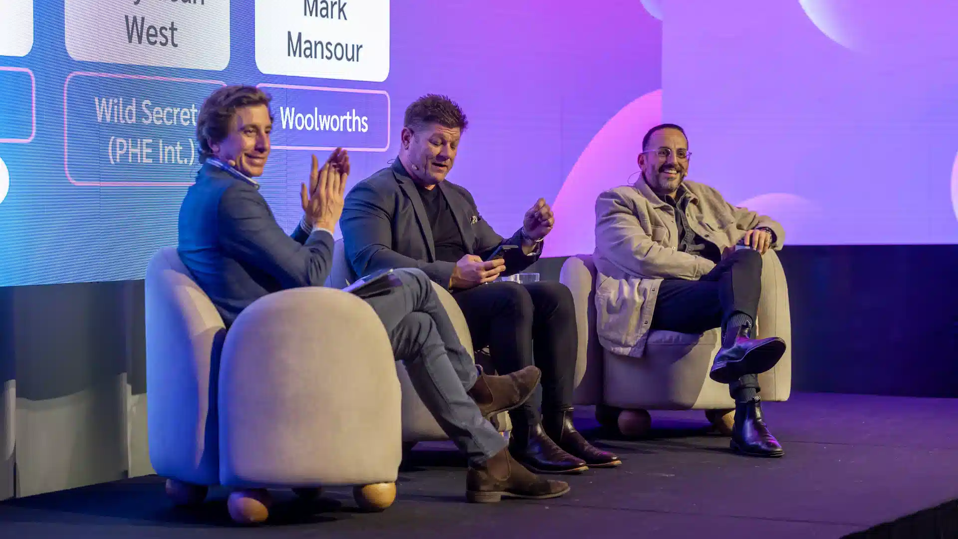 Three men seated on chairs engaged in discussion at a conference setting.