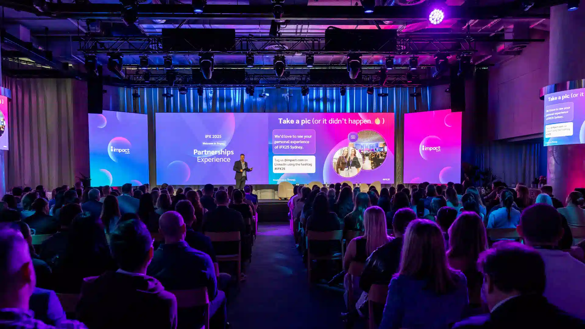 A large audience attentively watches a speaker presenting on stage, with bright lights illuminating the scene.