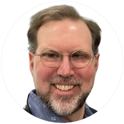 A man with light brown hair and glasses, wearing a blue shirt, situated against a neutral background in a circular frame.