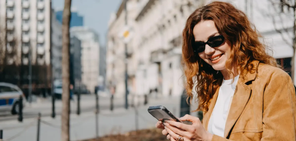 A woman wearing sunglasses is focused on her phone, appearing engaged with its screen.