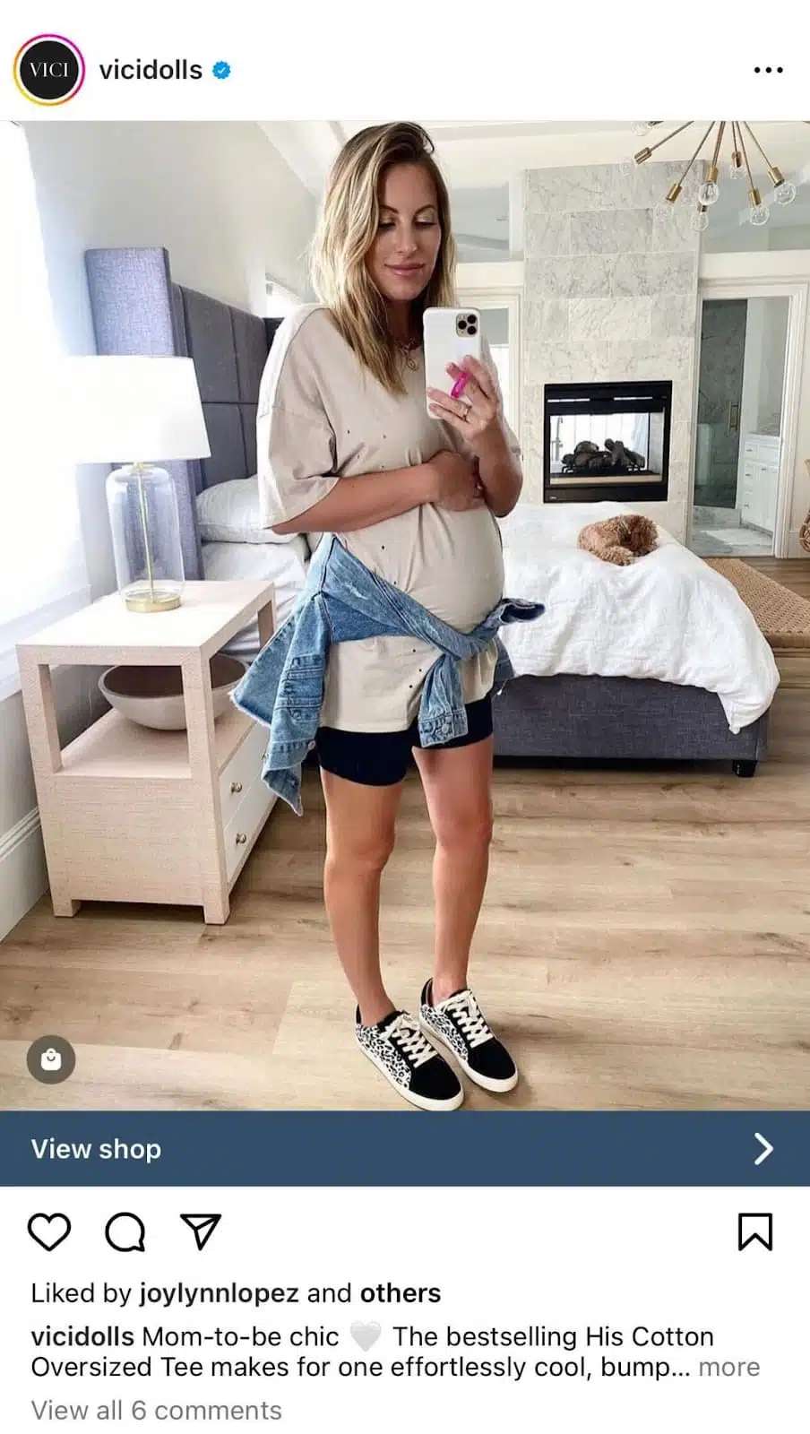 A pregnant woman stands in a stylish, light-filled living room wearing an oversized beige tee, black shorts, and casual sneakers.