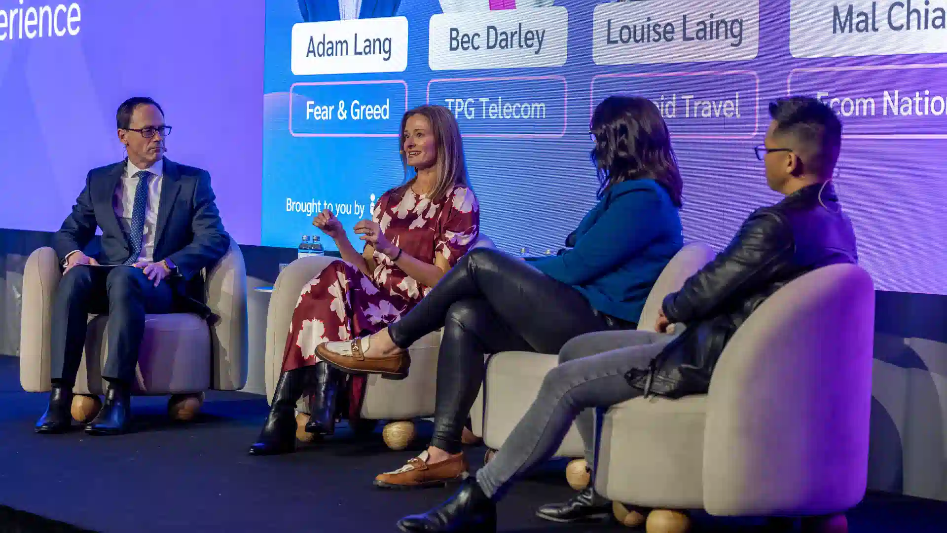 A panel discussion featuring four speakers seated on stage, discussing various topics, with vibrant background visuals.