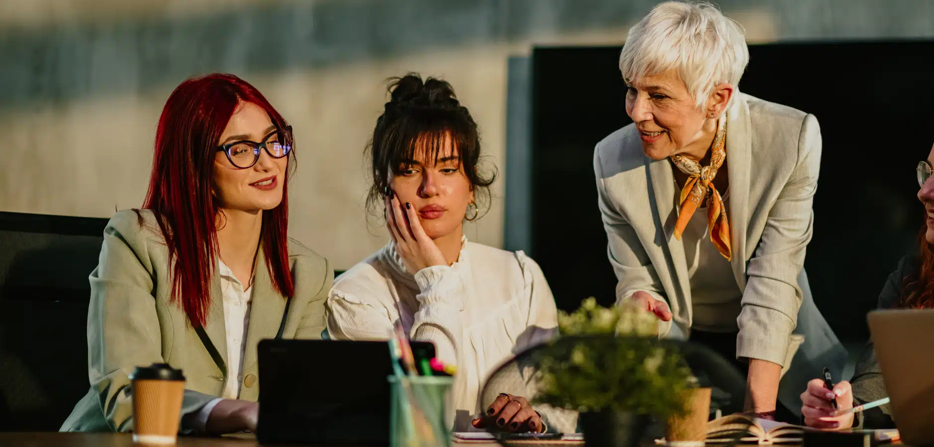 A group of professionally dressed women engaged in a work discussion at a modern office table, with laptops and stationery in view.