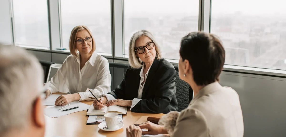Three businesswomen engaged in discussion at a conference table, with documents and laptops in front of them.