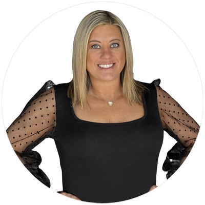 A woman in a black top with sheer dotted sleeves poses confidently, accessorized with a heart-shaped necklace.