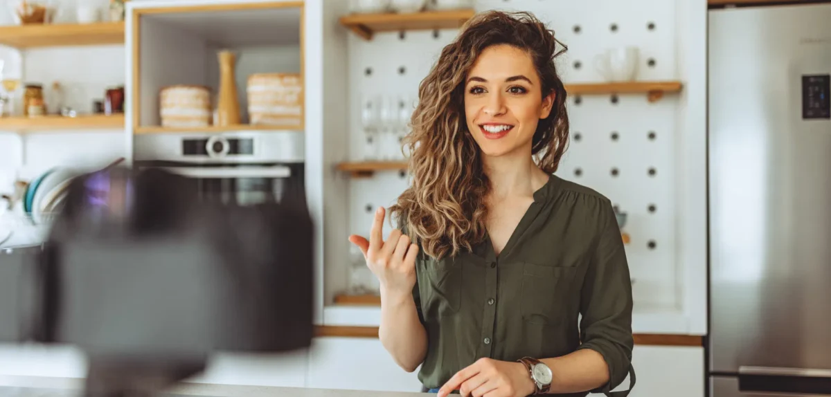A woman stands in a kitchen, facing a camera, with various cooking utensils visible around her.