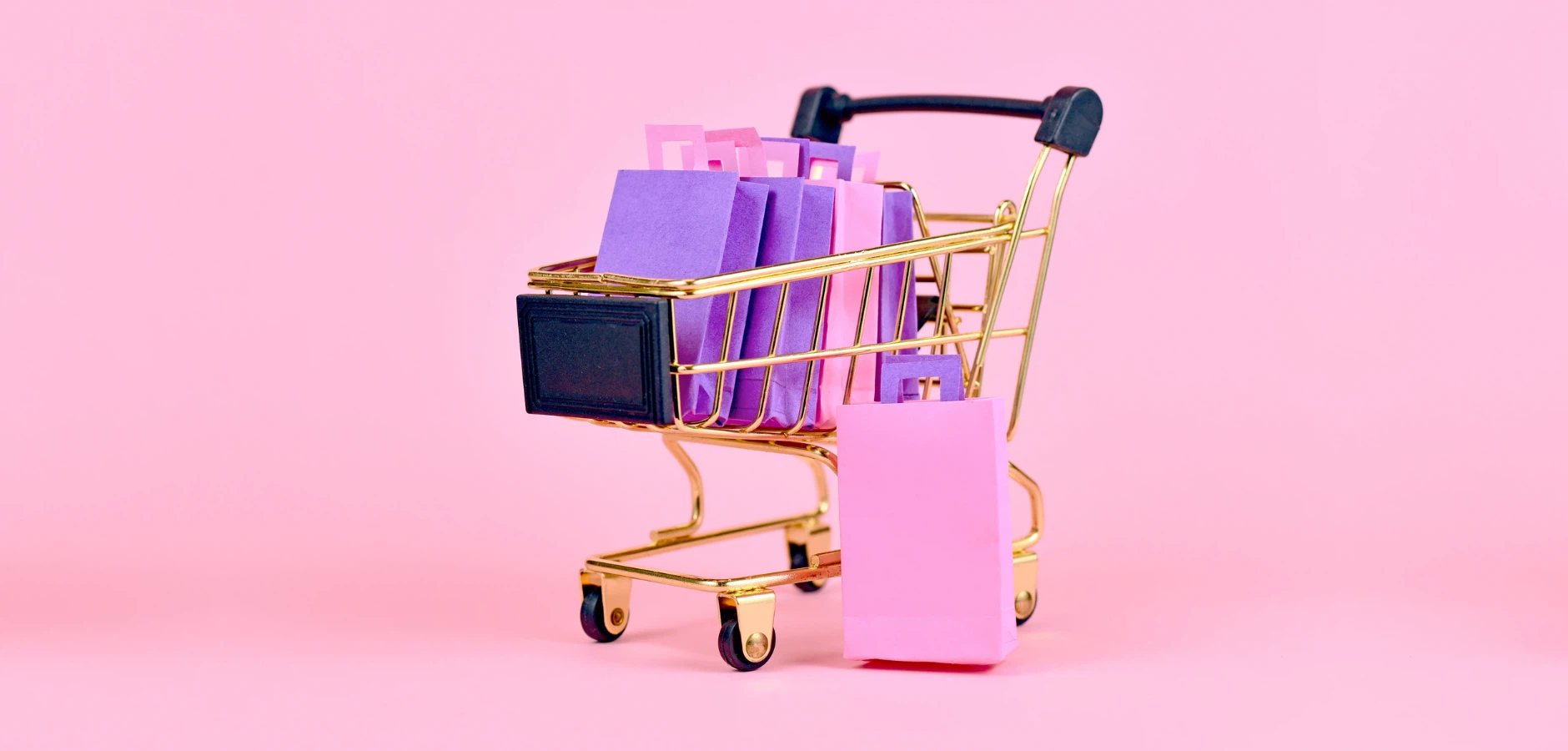 A gold shopping cart filled with purple and pink paper bags against a bright pink background.