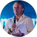 A speaker in a light-colored shirt holds a microphone, gesturing as they engage with an audience in a vibrant setting.