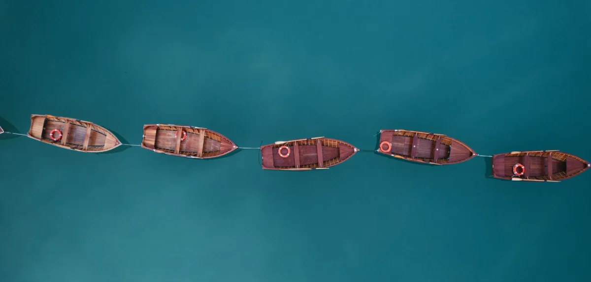 Aerial view of five wooden boats lined up on calm, turquoise water, each with a life ring.