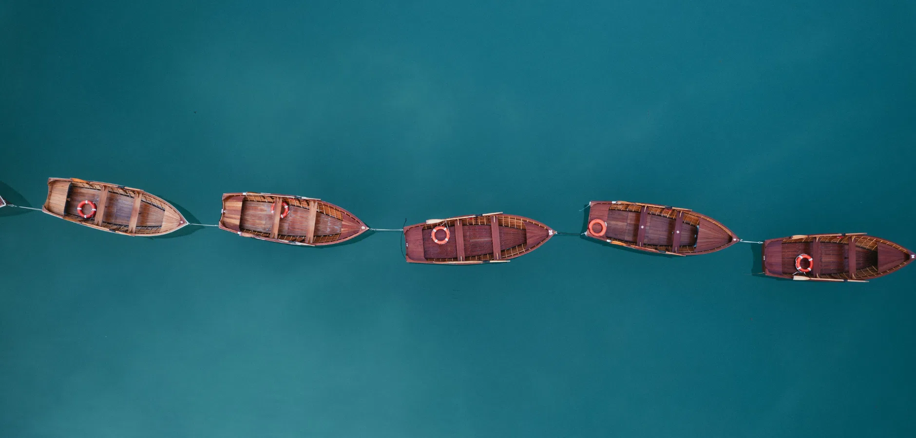 Aerial view of five wooden boats lined up on calm, turquoise water, each with a life ring.