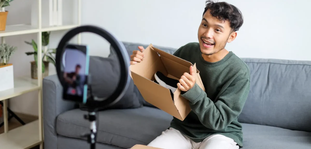 A person sits on a couch, holding an open cardboard box with shoes inside, while filming a video with a smartphone on a tripod.