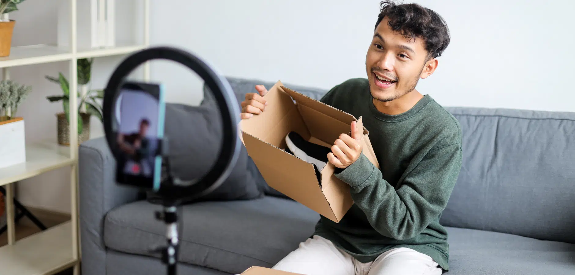 A person sits on a couch, holding an open cardboard box with shoes inside, while filming a video with a smartphone on a tripod.
