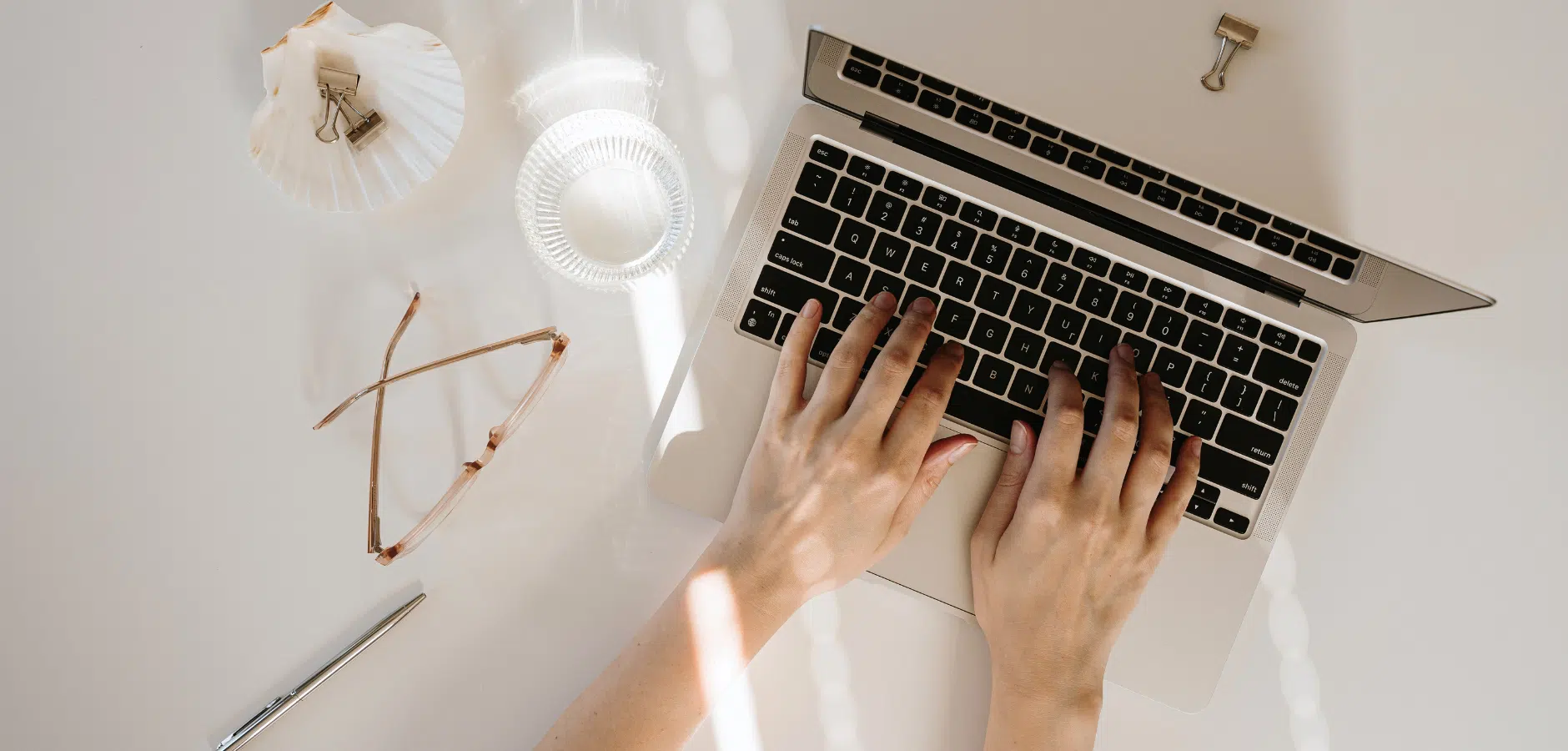 A flat lay of a workspace featuring a laptop, glasses, a pen, and a shell, illuminated by soft sunlight.